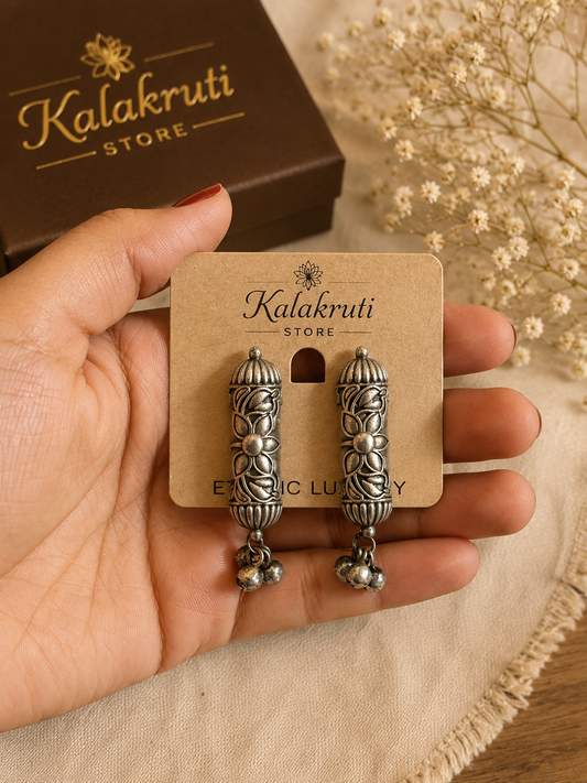 A hand holds a pair of ornate silver-toned earrings on a tan display card branded Kalakruti Store. The earrings feature intricate filigree detailing and dangling bell charms.
