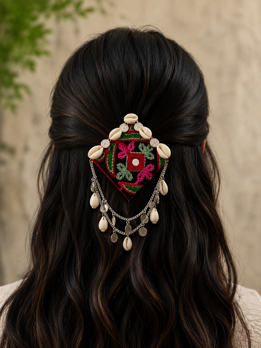 A woman viewed from the back with long, wavy dark brown hair, wearing a decorative embroidered hair accessory with shell-like beads, silver-toned chains, and colorful floral embroidery in red, green, and pink.