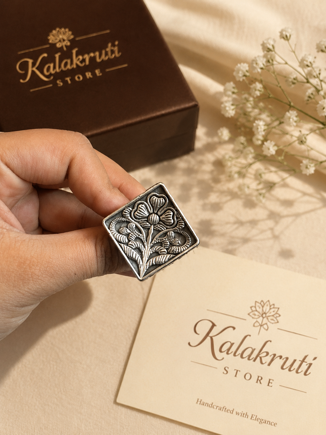 A hand holds a square silver-toned metal stamp with an embossed floral design, with a brown Kalakruti Store box and a cream-colored branded card in the background on a beige fabric surface with small white flowers.