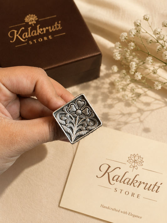 A hand holds a square silver-toned metal stamp with an embossed floral design, with a brown Kalakruti Store box and a cream-colored branded card in the background on a beige fabric surface with small white flowers.