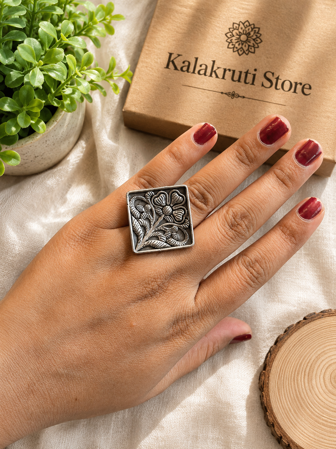 A silver-toned square ring with an embossed floral pattern worn on a person's hand, styled with a kraft paper box labeled 'Kalakruti Store', a small green plant, and a wood slice on a cream fabric background.