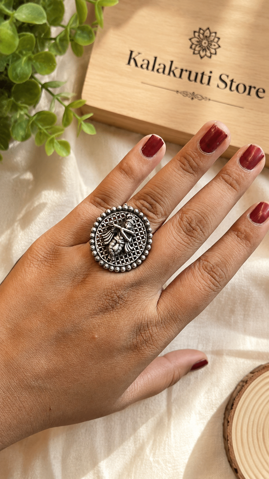 A silver-toned ring with an intricate bird design, worn on a hand with dark red nail polish. The background features a wooden box labeled 'Kalakruti Store' and green leaves.