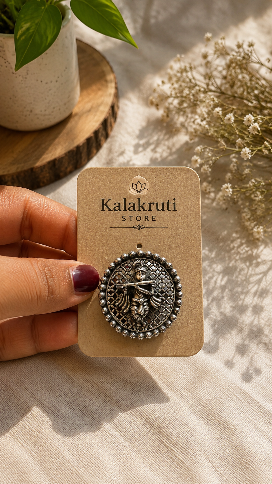 A hand with dark burgundy nail polish holds a round silver-toned pendant on a tan display card. The card is branded 'Kalakruti Store' and features a small illustration of a woman playing a stringed instrument.