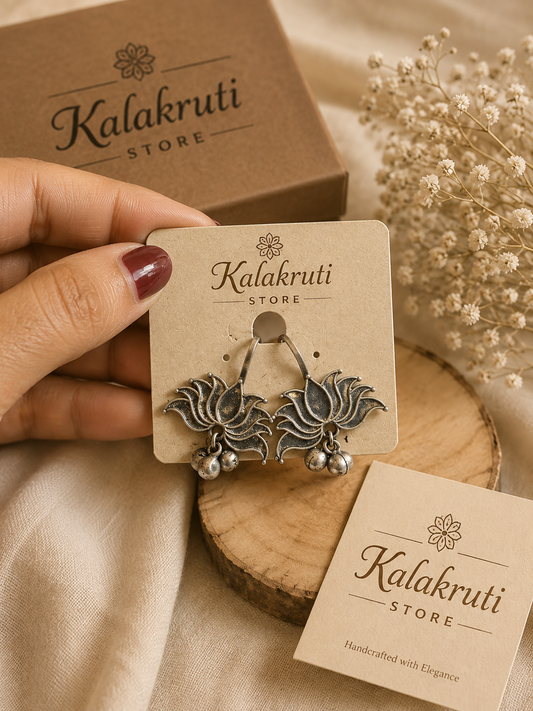 A hand with dark red nail polish holds a pair of silver-toned lotus flower earrings on a beige display card labeled 'Kalakruti STORE'. The scene includes a brown kraft box, dried baby's breath flowers, and a wooden slice on a beige fabric background.