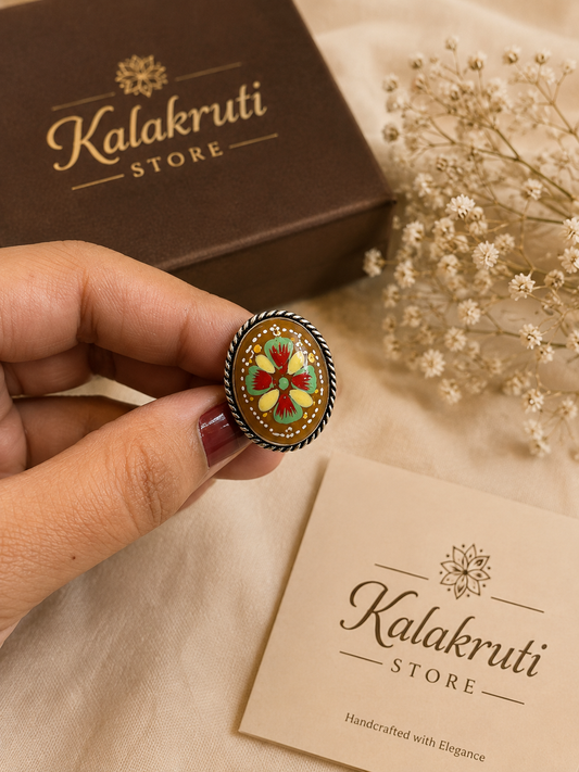 A hand holds a round enamel pin with a floral design in red, yellow, and green on a dark metal background. A brown Kalakruti Store box is in the background, along with a beige card and dried flowers on a light fabric surface.
