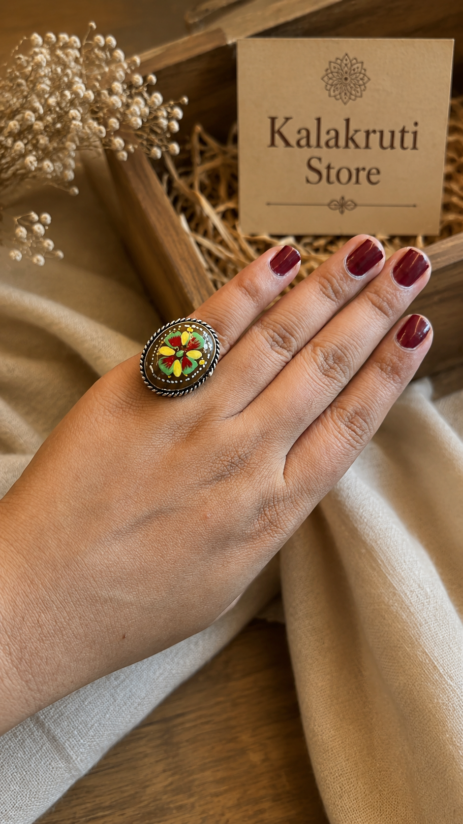 A hand with dark red painted nails wears a large oval ring featuring a colorful floral mandala design in yellow, green, and red on a dark metal background. The hand is resting on beige fabric next to dried flowers and a card for 'Kalakruti Store'.