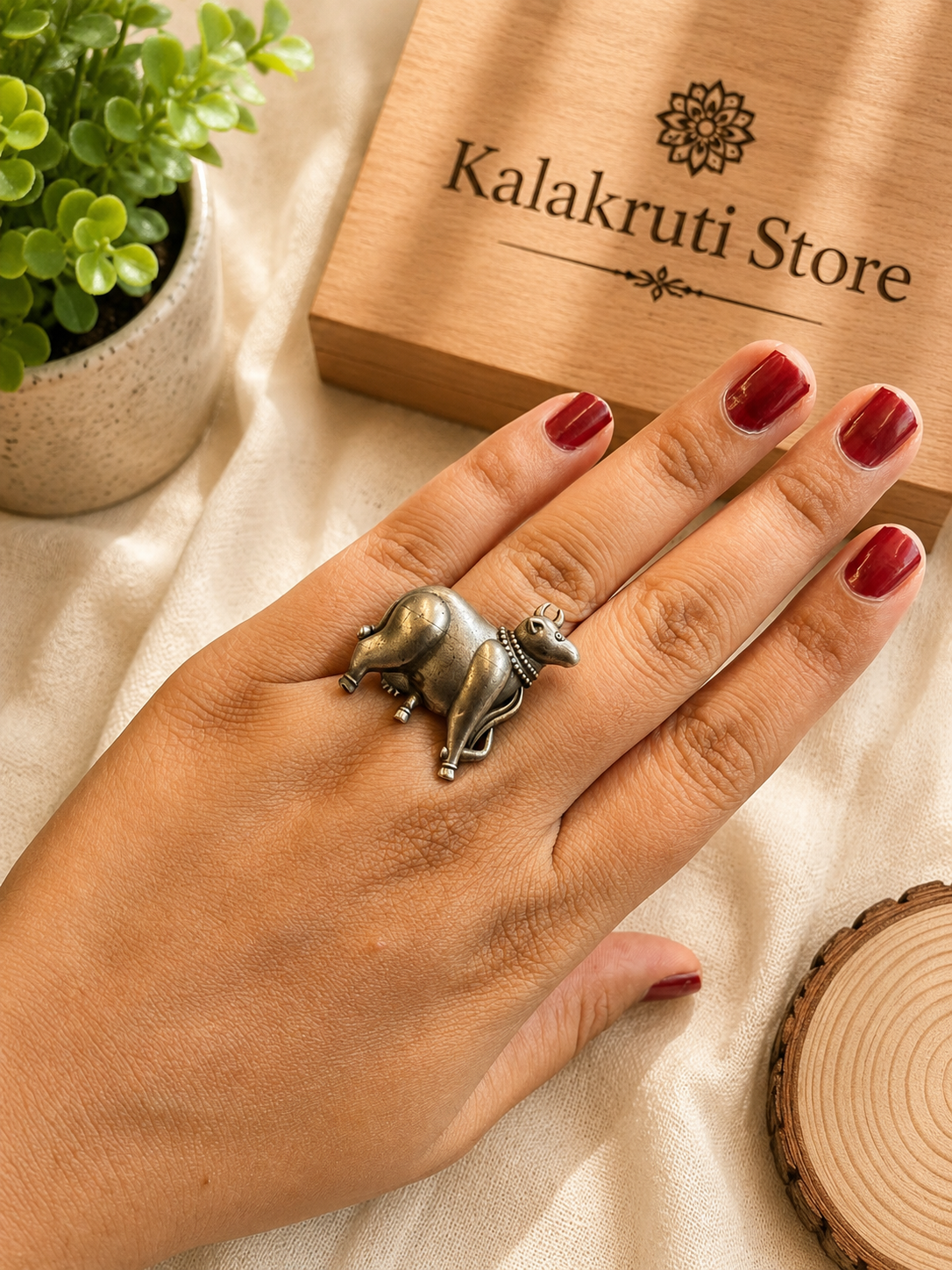 A hand with red nail polish wearing a silver-toned cow ring, resting on a beige fabric surface next to a wooden box branded Kalakruti Store.