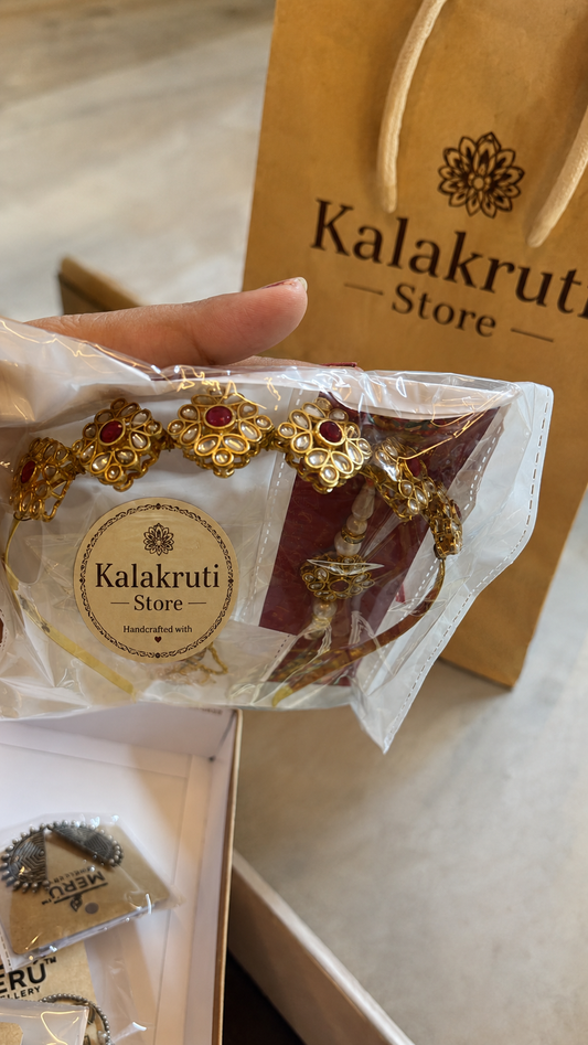 A hand holds a packaged Kalakruti Store gold-toned bracelet with red stone accents in front of a brown Kalakruti Store shopping bag on a light surface.