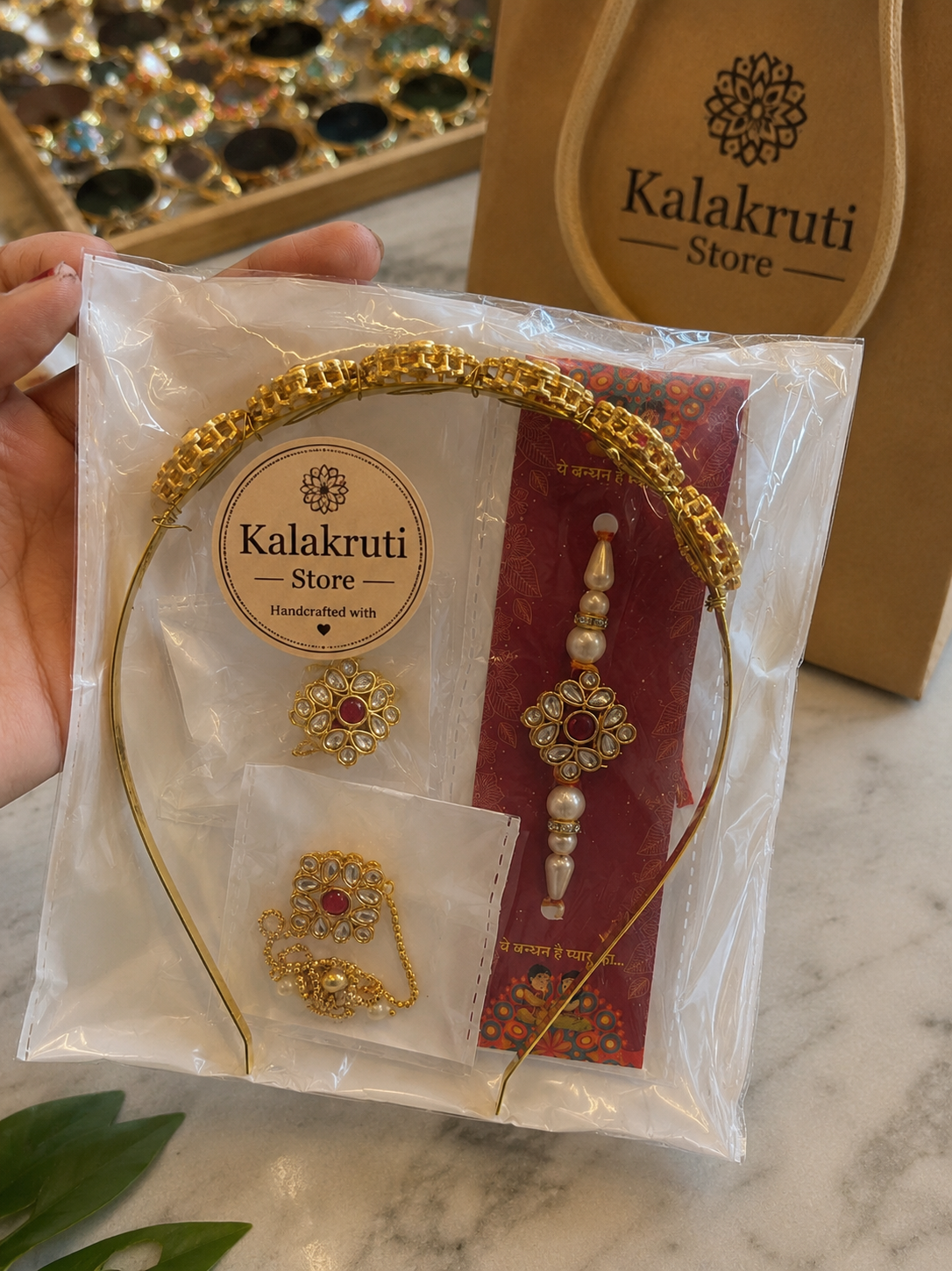 A hand holds a clear plastic bag containing a gold-toned jewelry set with a decorative headband, earrings, and a red card insert. The packaging shows the brand name 'Kalakruti Store', and a matching brown paper bag is visible in the background on a marble surface.