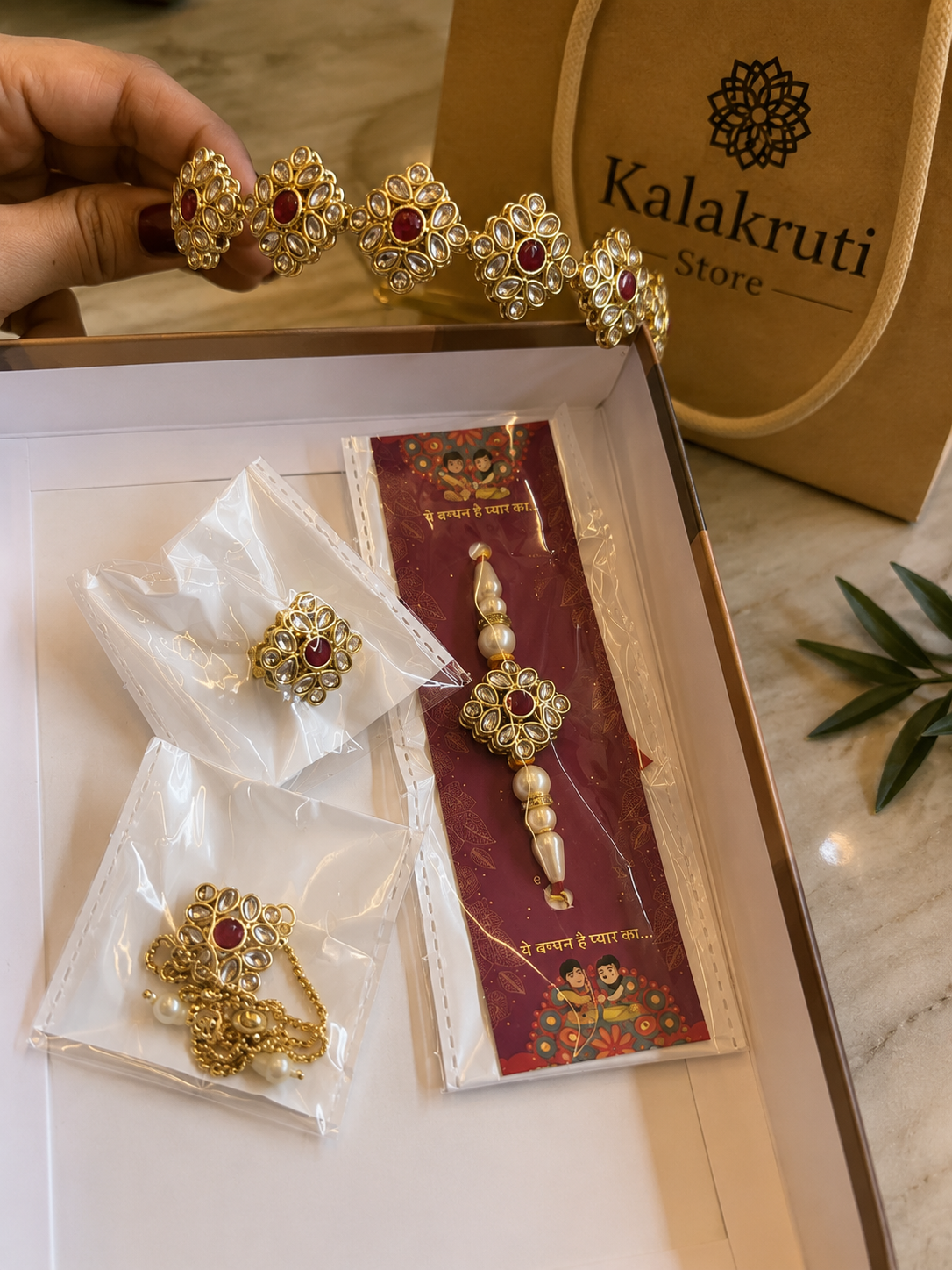 Gold-toned traditional Indian jewelry set with red stones and pearl-like accents, displayed in an open box alongside branded Kalakruti Store packaging and a gift bag.