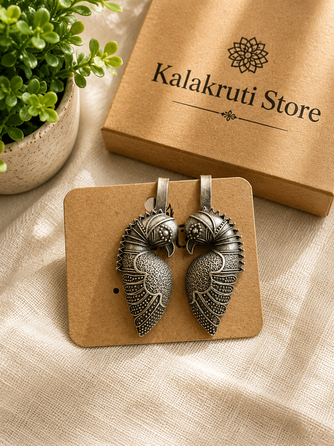 A pair of ornate silver-toned wing-shaped earrings displayed on a small kraft card, with a kraft box labeled 'Kalakruti Store' and a potted green plant in the background on a beige fabric surface.