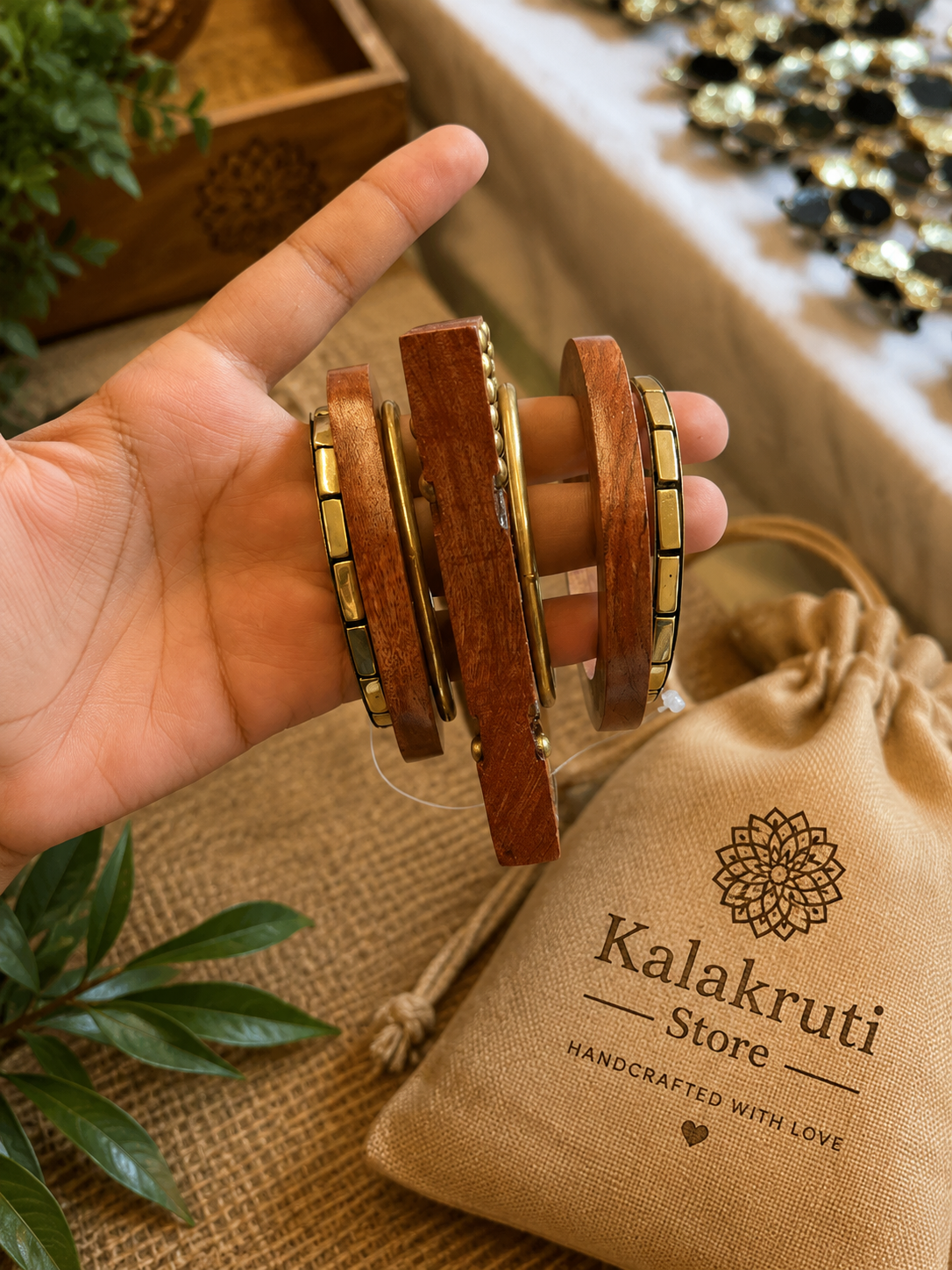 A hand holds a stack of wooden bangles with gold-toned metal accents. A beige drawstring pouch printed with the brand name 'Kalakruti Store' sits nearby on a burlap surface, with decorative plants and jewelry in the background.