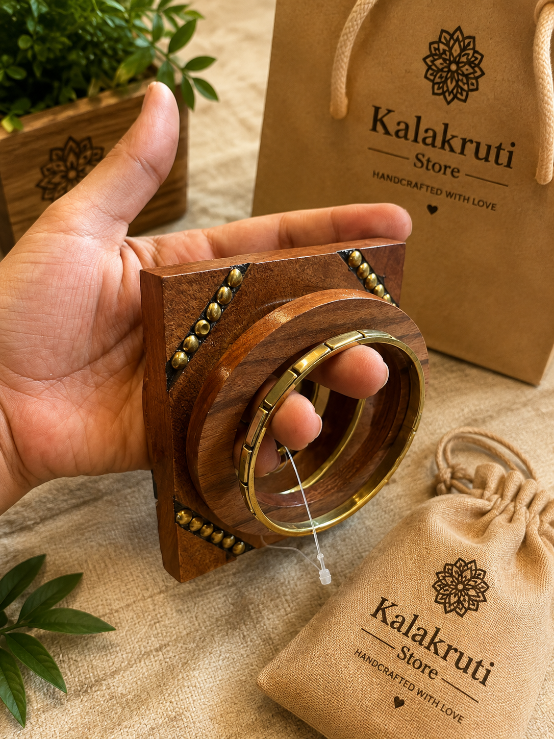 A hand holds a wooden jewelry display stand showcasing a gold-toned bangle, with branded kraft paper bags and a drawstring pouch in the background on a beige fabric surface.