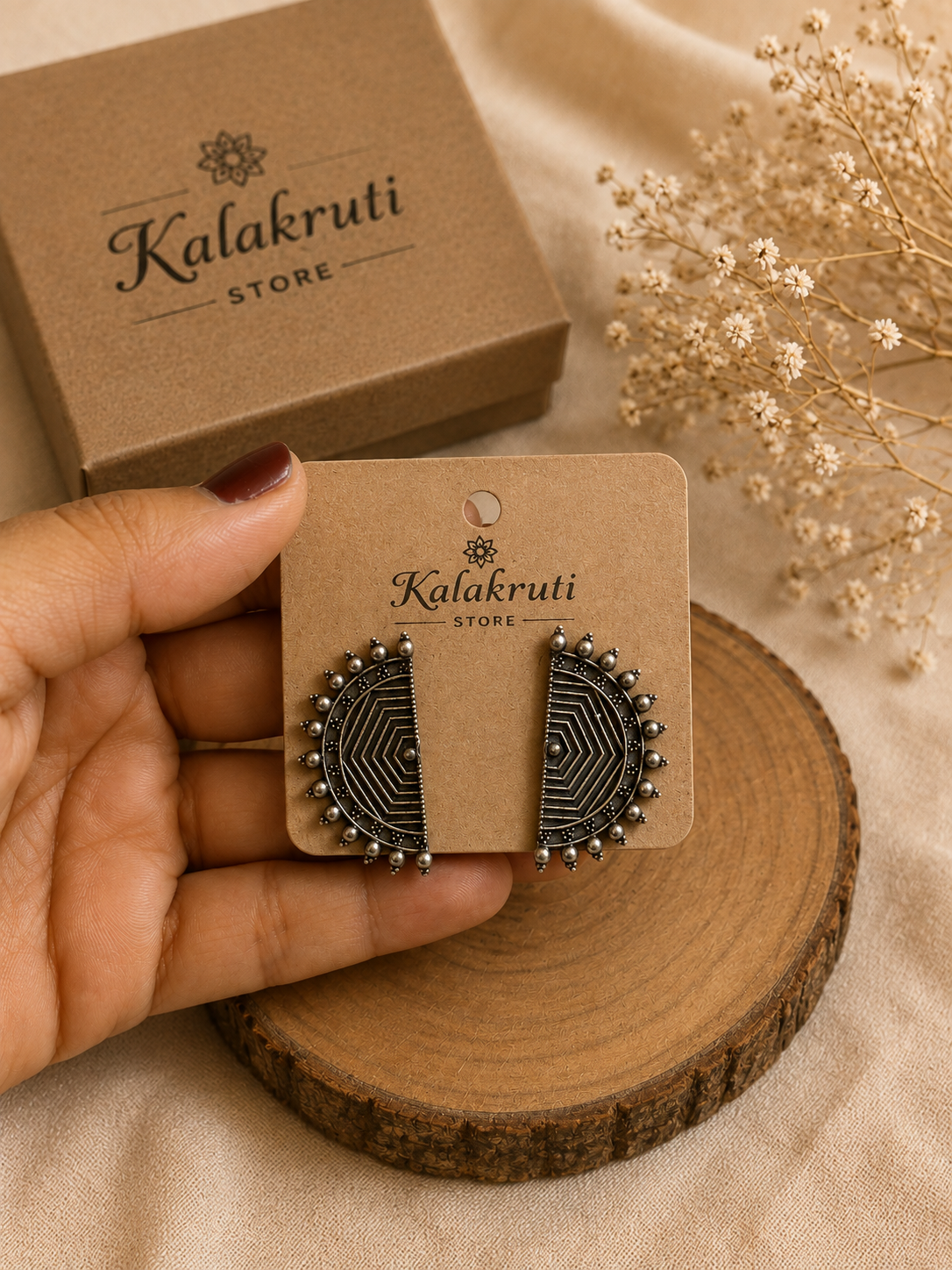 A hand holds a pair of black geometric stud earrings on a brown display card branded Kalakruti Store, with a matching box and dried flowers in the background on a beige fabric surface.